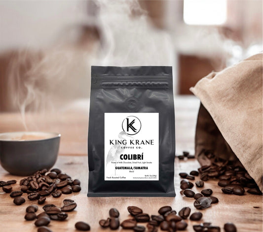 A package of King Krane Colibrí coffee with a background featuring a cup of coffee and loose coffee beans on a table.