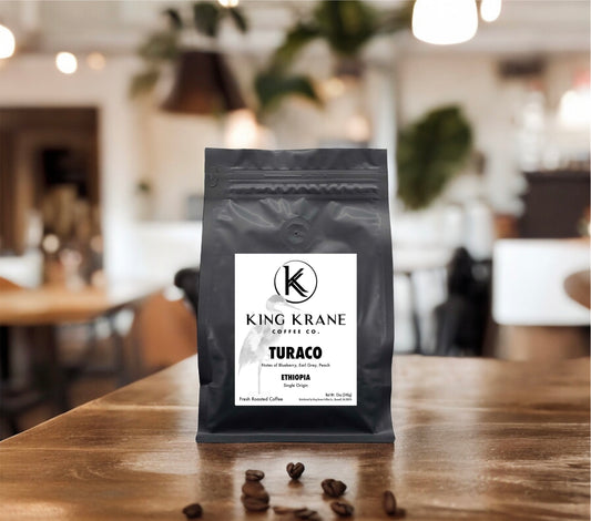 A packaged bag of King Krane Turaco coffee beans on a wooden surface, with a label showing the brand and coffee name, and a background featuring a blurred cafe setting.